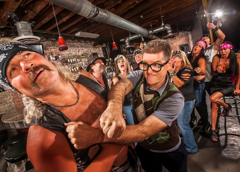 Group of diverse individuals in a bar setting, with a man in a leather vest and bandana playfully interacting with another man in glasses, surrounded by others laughing and enjoying drinks, highlighting a lively atmosphere related to social gatherings and potential liquor liability discussions.