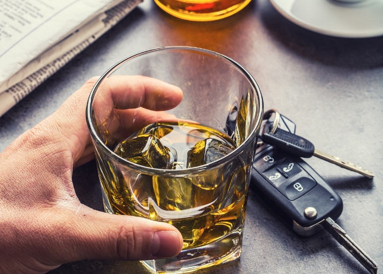 Hand holding a glass of whiskey with ice, keys on a table, and a newspaper, highlighting alcohol consumption and potential liability under New Jersey liquor laws.