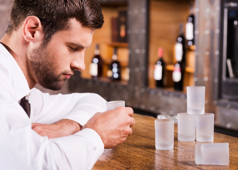 Man in a white shirt thoughtfully holding a glass, surrounded by frosted shot glasses on a bar, with liquor bottles visible in the background, relevant to New York Dram Shop Act discussions.