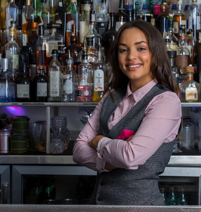 Smiling bartender in a vest standing confidently behind a bar stocked with various liquor bottles, emphasizing responsible alcohol service in hospitality.