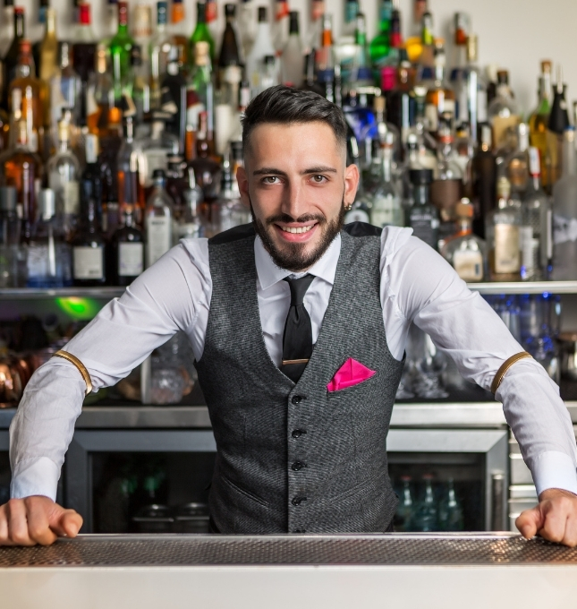 Bartender smiling confidently at a bar with a display of various liquor bottles, representing responsible alcohol service and management in hospitality settings.