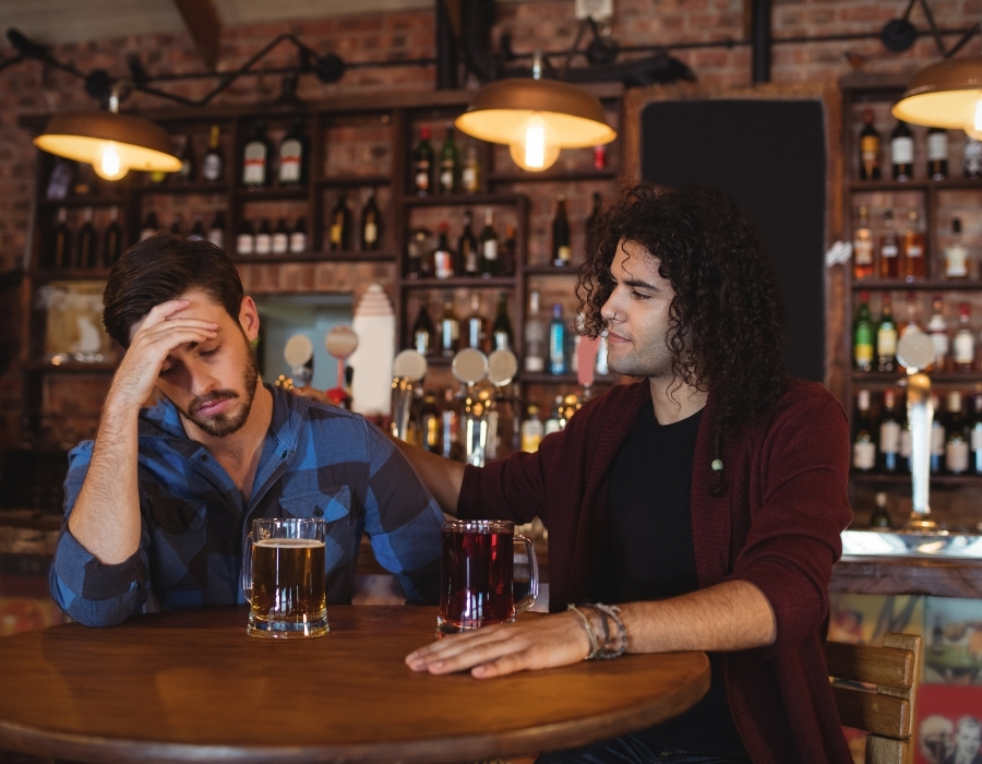 Two men sitting at a bar table, one looking distressed with a hand on his forehead and a glass of beer in front of him, while the other offers support with a hand on his shoulder and a drink beside him, illustrating the importance of recognizing and addressing alcohol-related issues.
