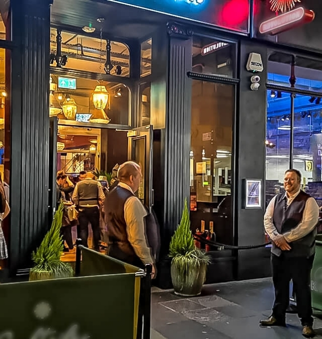 Restaurant entrance with staff in vests, patrons entering, highlighting hospitality and alcohol service standards.