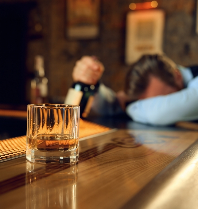 Man with head down on bar, holding a bottle, and a glass of whiskey in foreground, illustrating issues of alcohol service and overservice in bars and restaurants.